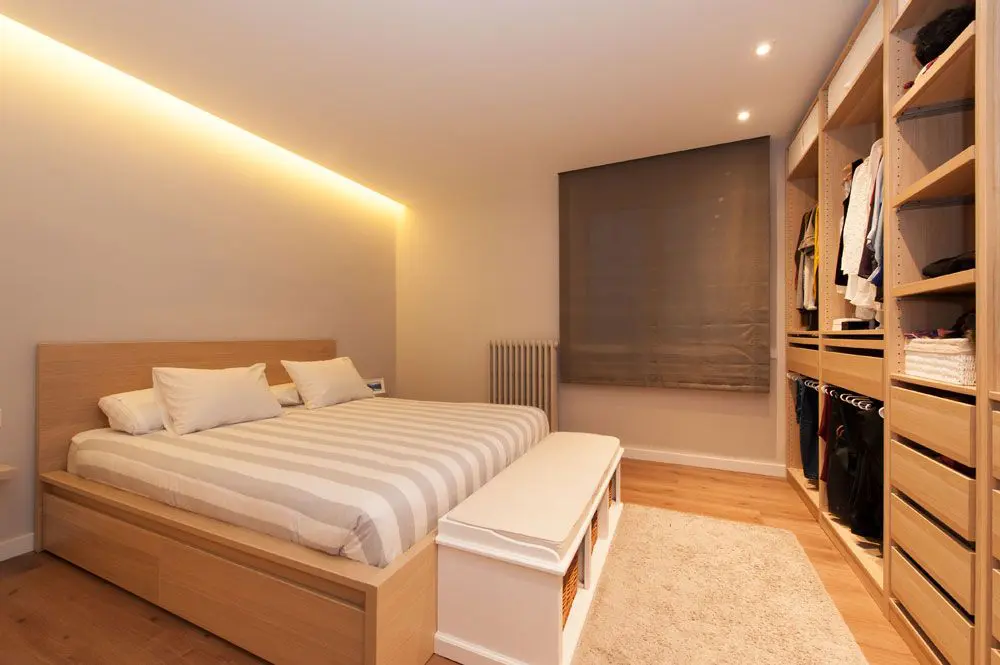 Vestidor de dormitori amb un armari obert al llarg de la paret de davant del llit. Amb banc i catifa. Projecte Sincro a Barcelona.