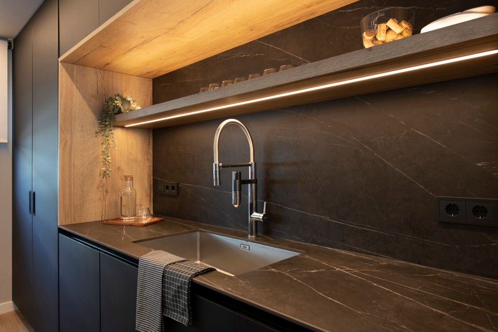 Hueco mueble con la zona de fregadero cocina con tonos oscuros combinado con madera. Con tiras led iluminación.
