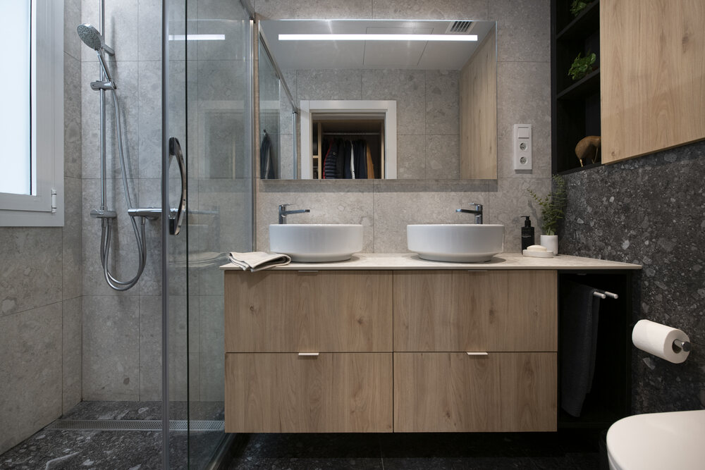 Baño con ducha con mueble suspendido de madera con doble pica sobre encimera. Baldosas terrazo en tonos grises claros y oscuros.