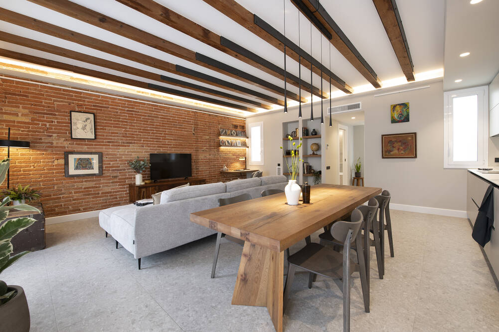 Salón comedor con pared ladrillo visto y vigas en techo de madera. Suelo de terrazo gris.