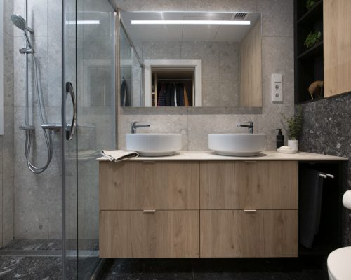Baño con ducha con mueble suspendido de madera con doble pica sobre encimera. Baldosas terrazo en tonos grises claros y oscuros.